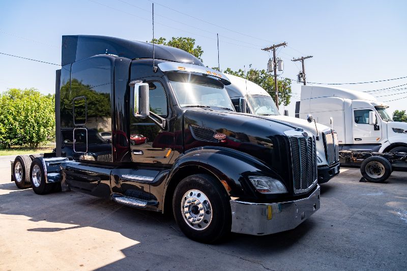 Shiny black Peterbilt semi-truck parked beside white trucks in a sunny lot, with clear skies and utility poles in background.
