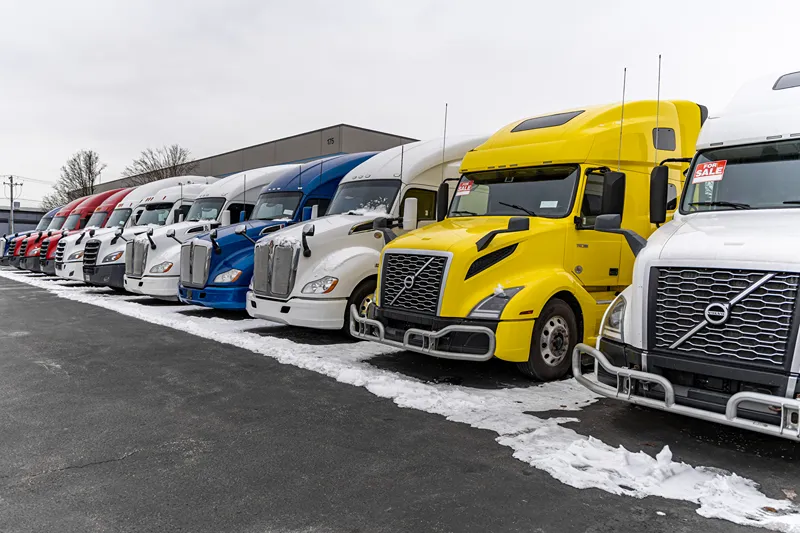 Heavy-duty truck sales lot with rows of semi-trucks parked in snow and “FOR SALE” signs displayed.