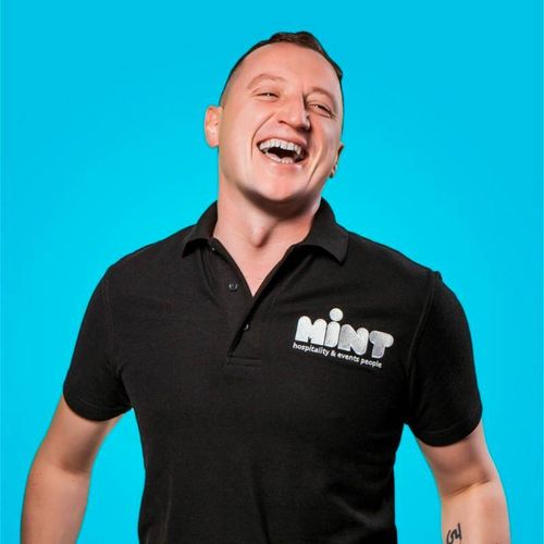 Smiling man wearing a black polo shirt with the logo 'Mint hospitality & events people' on a bright blue background.