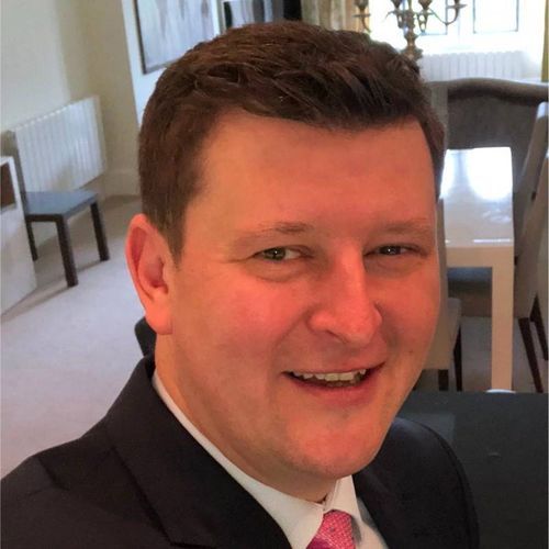 Smiling man with short brown hair wearing a dark suit jacket, white shirt, and pink tie in a dining room.