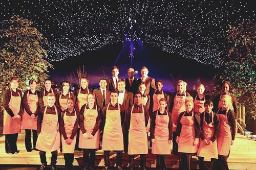 Group photo of 22 people, most wearing pink aprons over black shirts, standing on a stage with decorative trees and star-like lights above.