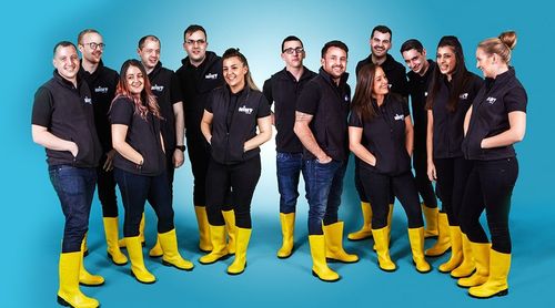 Group of people wearing black tops and bright yellow boots standing on a blue background, smiling and interacting.