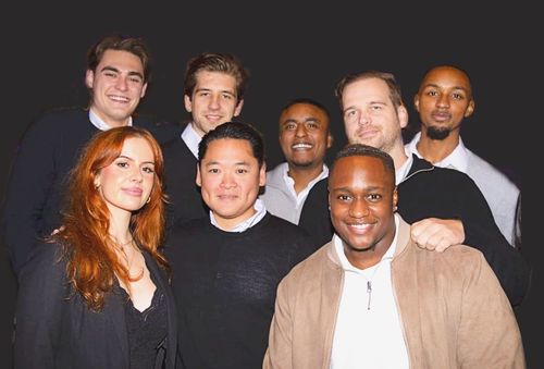 Group of seven diverse young adults smiling and posing against a dark background.