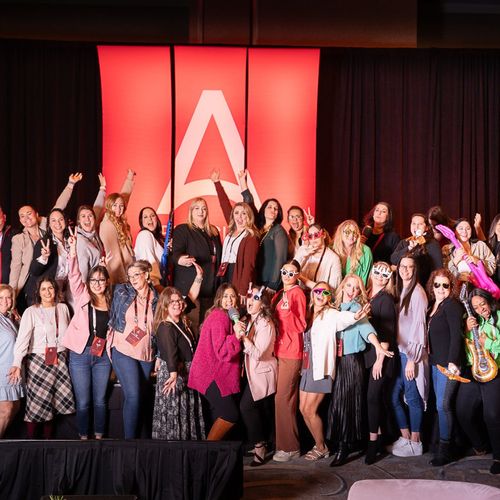 Group of diverse women posing energetically on stage in front of a large red backdrop with a white stylized letter A.