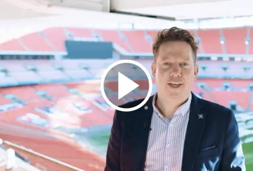 Man in a blazer speaking in an empty sports stadium with red seats.