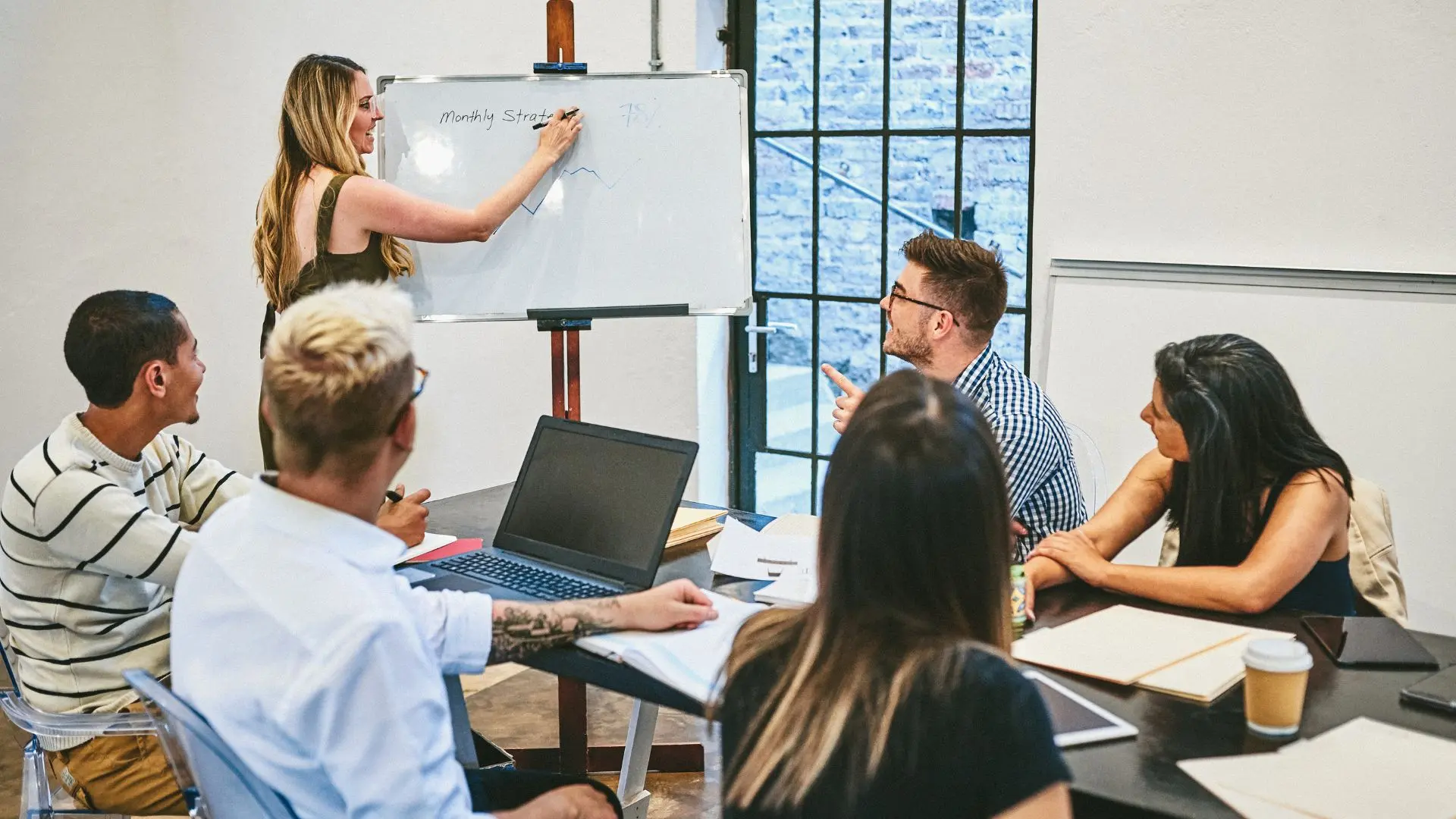 Eine Gruppe von Kolleg:innen sitzt in einem modernen Besprechungszimmer um einen Tisch mit Laptops und Notizblöcken. Eine Frau steht an einem Whiteboard und zeichnet ein Liniendiagramm unter der Überschrift "Monthly Strategy", während das Team aufmerksam zuhört und interaktiv diskutiert. Das Bild symbolisiert strategische Personalentwicklung, kontinuierliche Lernprozesse im Unternehmen, agile Workshops und das gemeinsame Schließen von Kompetenzlücken in modernen Führungsteams.