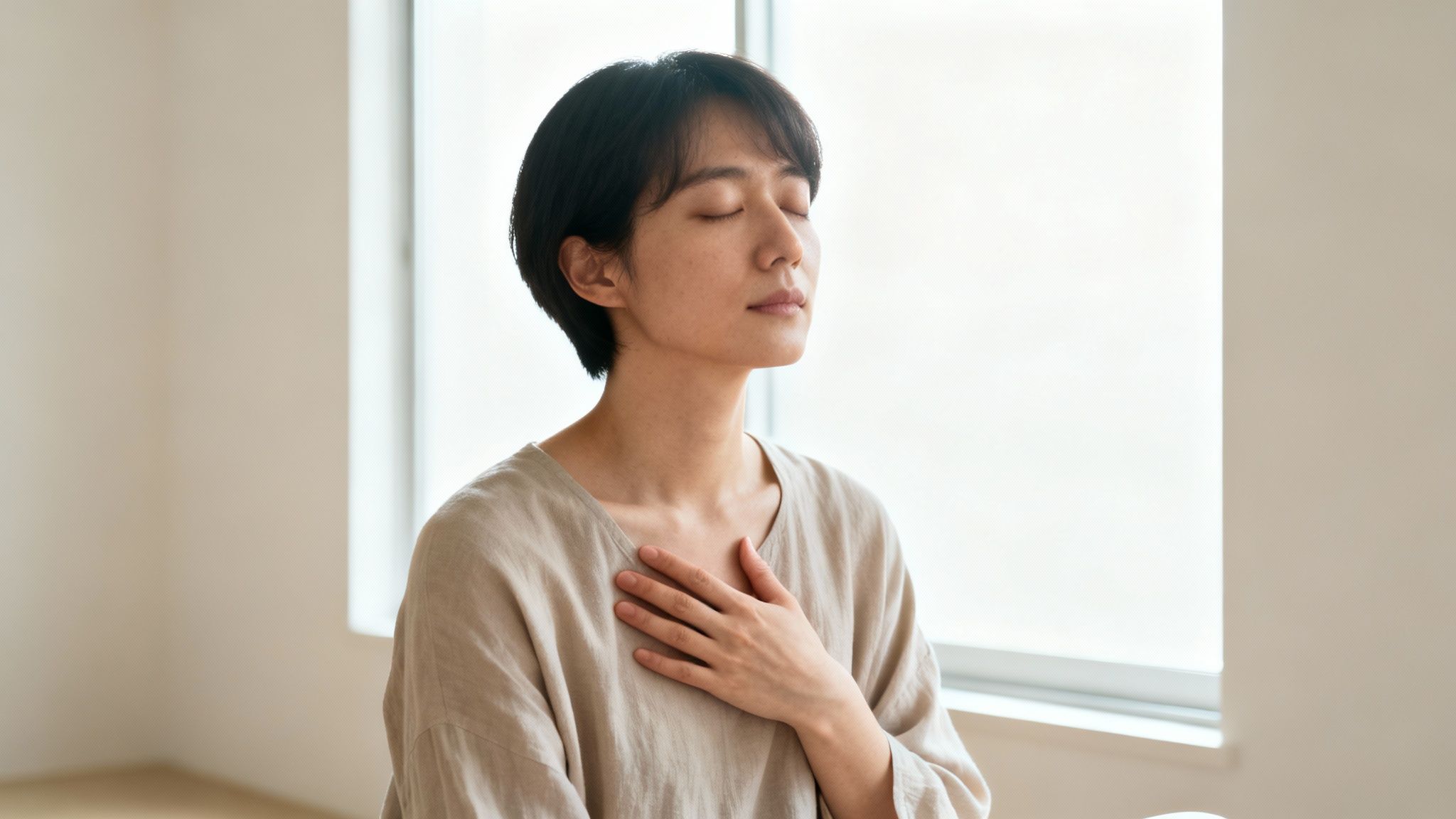 A serene person with short hair meditates with eyes closed, hand on chest, in a bright room.
