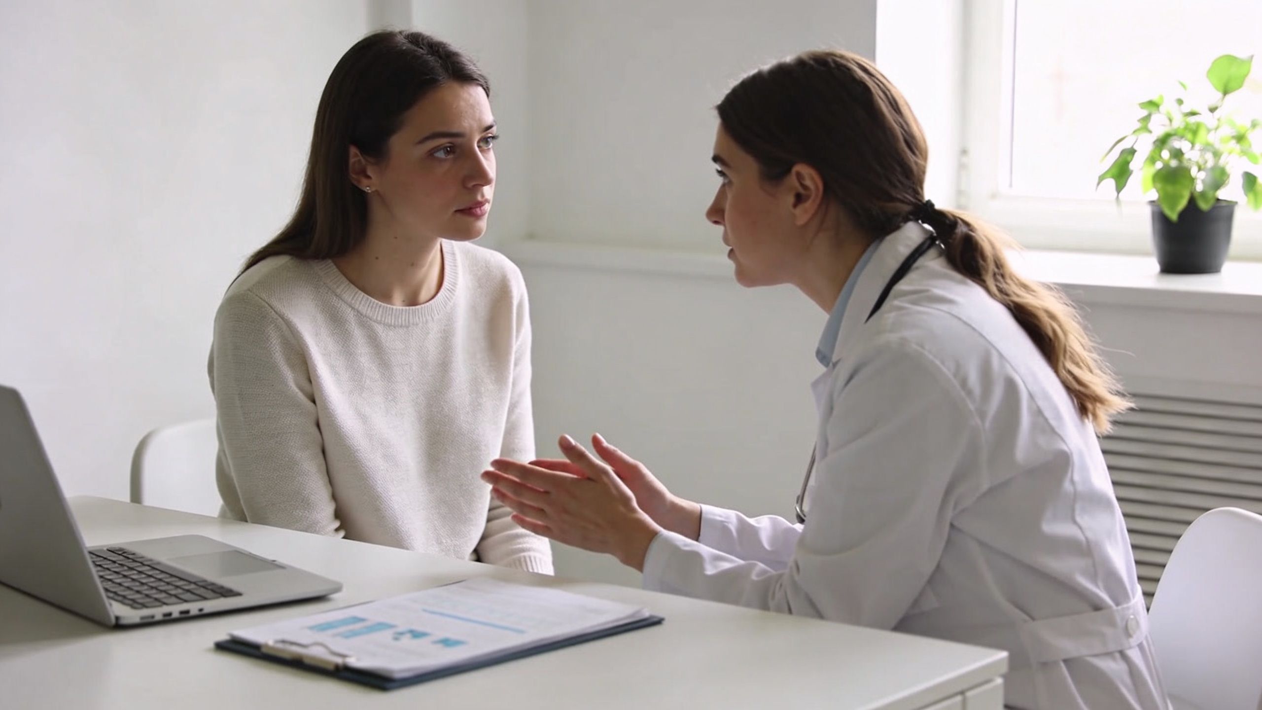 A female doctor in a white coat consulting with a female patient in a medical office.
