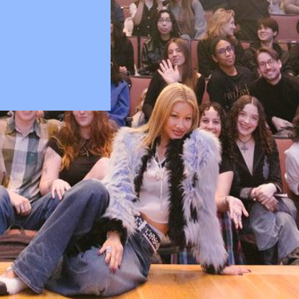 Yoon Ahn, in a blue and black shaggy faux-fur jacket, sits on the edge of the stage waving to the camera, with FIT students filling the tiered seating behind.