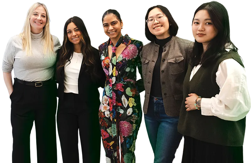 A diverse group of five women standing side by side, smiling at the camera, dressed in a mix of business and casual attire including blazers, a floral dress, and layered outfits, against a neutral background.