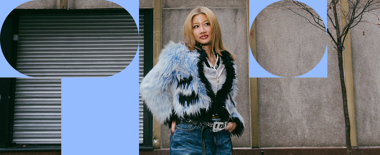 Yoon Ahn, co-founder and creative director of AMBUSH, in a light blue and black shaggy faux-fur jacket, white top and blue jeans, standing against a concrete wall and metal shutter door, framed by large pale blue graphic shapes.