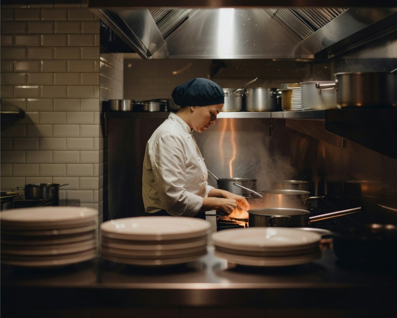 A chef working at the stove in a busy restaurant kitchen, with stacks of plates in the foreground