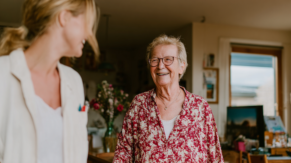A caregiver assists an elderly person near window plants.