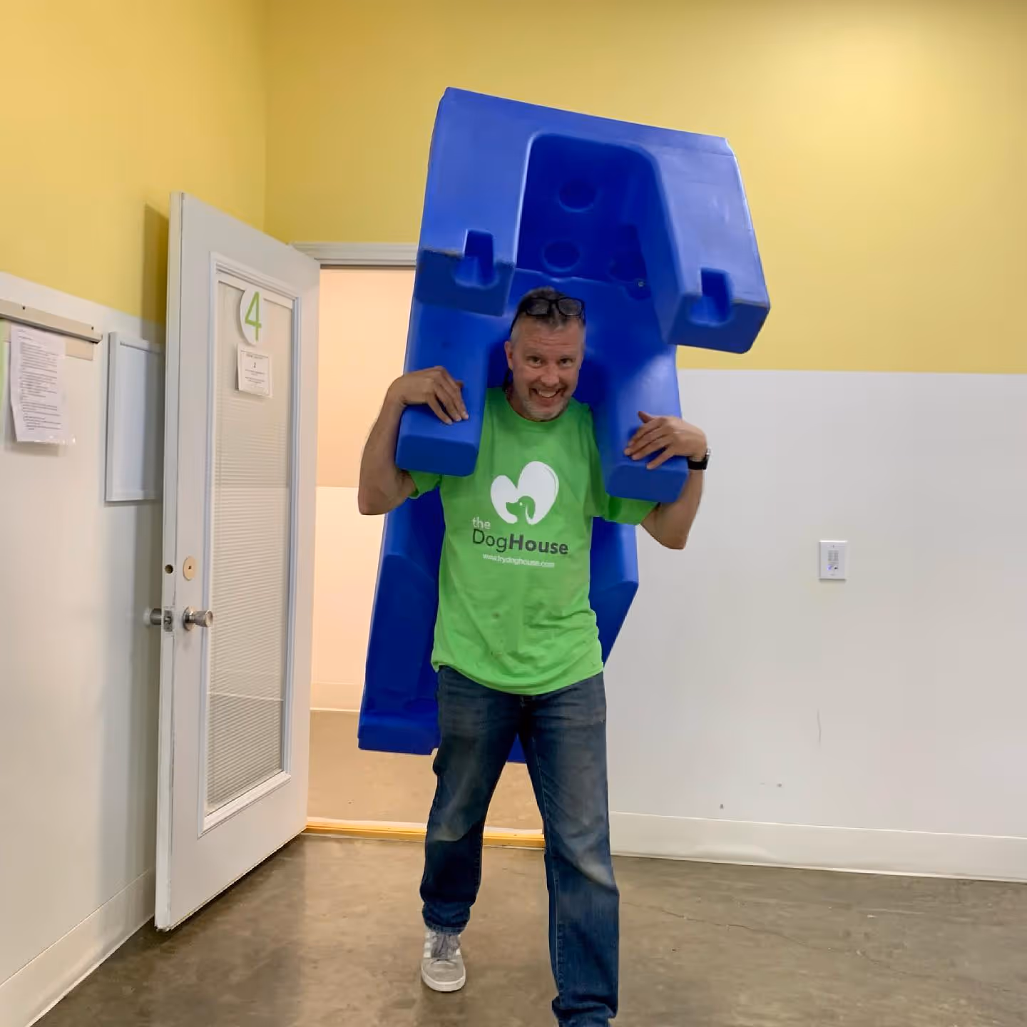 Man wearing green DogHouse t-shirt carrying a large blue plastic doghouse indoors.