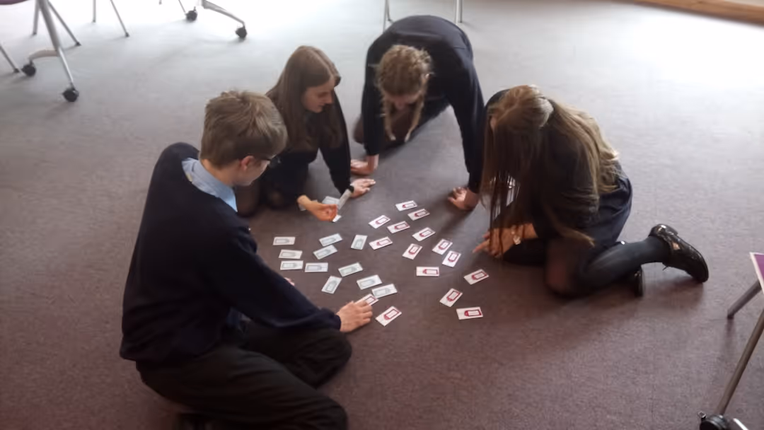 Four students in uniforms sitting on the floor playing a card game with cards spread out in a circle.