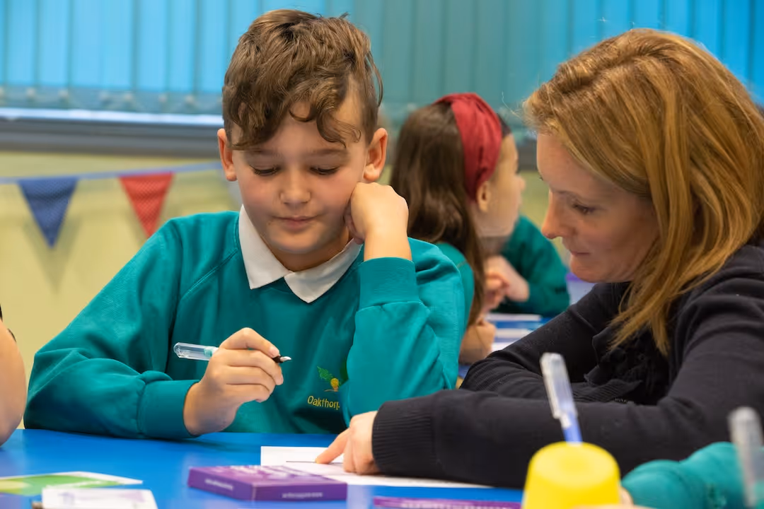 Teacher helping a student in green school uniform with writing at a blue classroom table.
