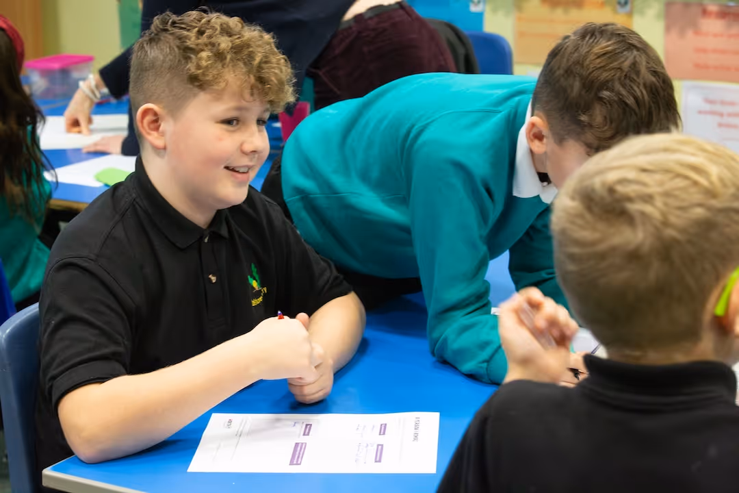 Smiling boy with curly hair sitting at a table with a worksheet in a classroom setting.