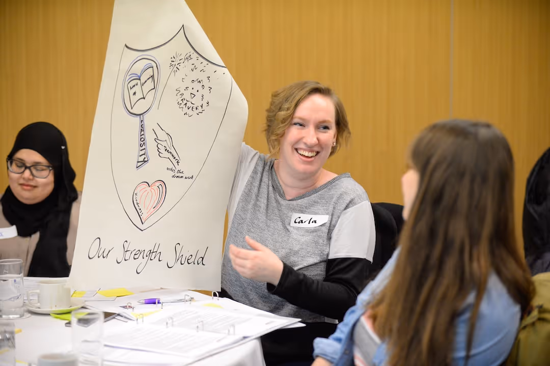 Teacher smiling and holding a large paper with drawings titled 'Our Strength Shield' at a meeting.