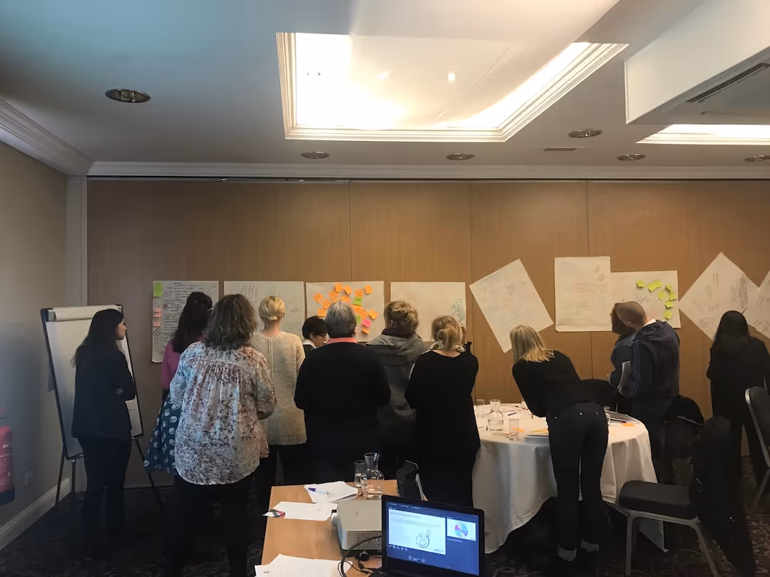 Group of teachers standing and looking at large sheets with notes and sticky notes on a wall in a meeting room.