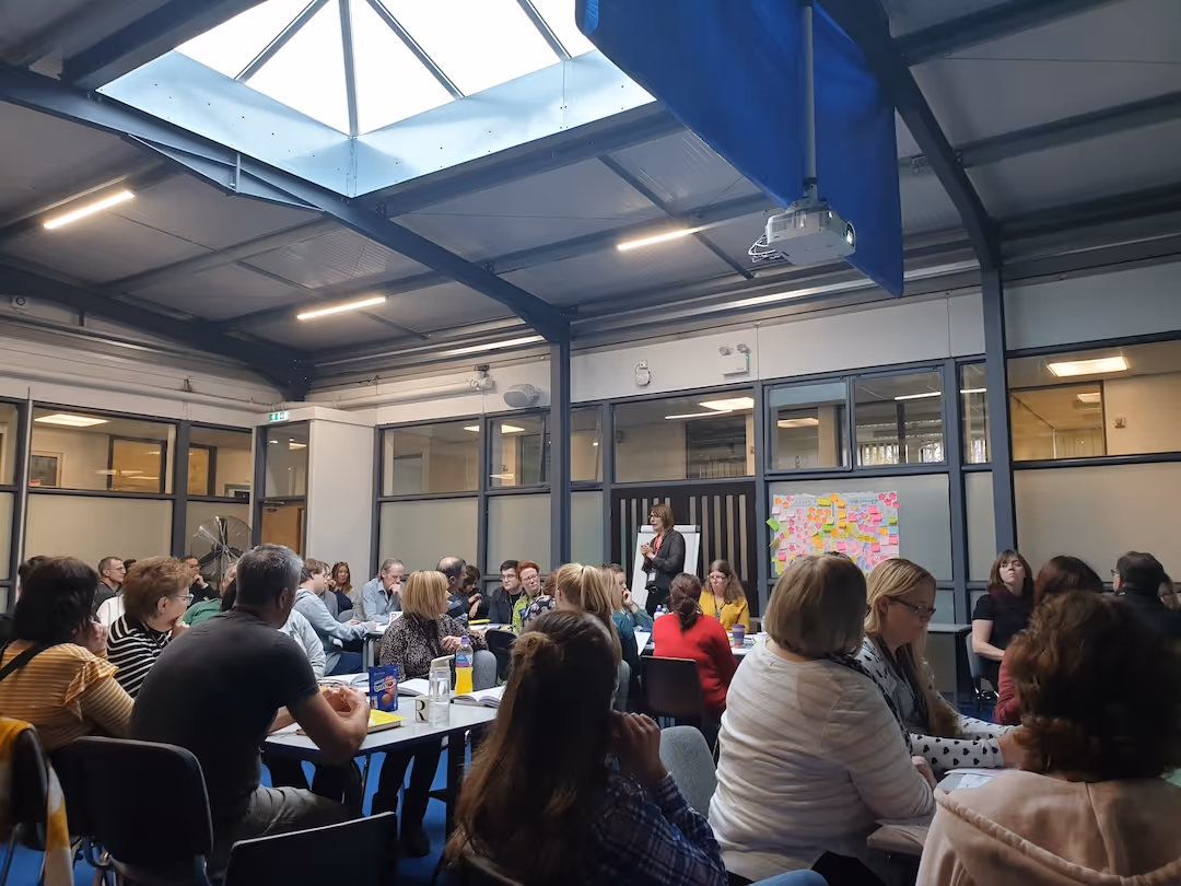 Group of teachers at a workshop in a room listening to a standing speaker near a wall with sticky notes.