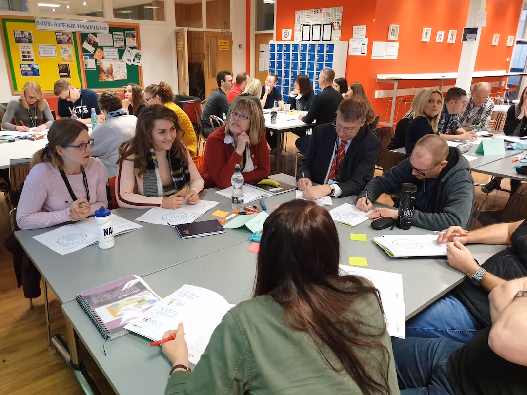 Teachers seated around tables in a workshop room engaged in writing and discussing documents.