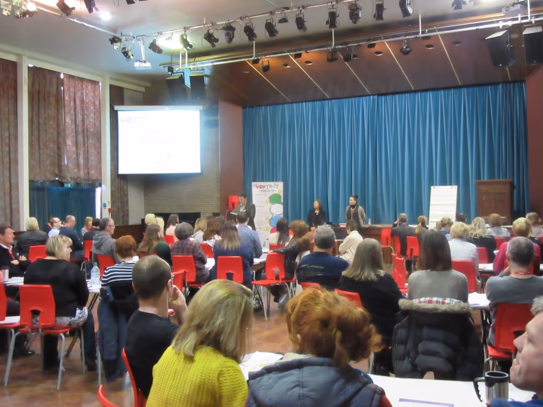 Group of people seated in red chairs attending a presentation in a large room with blue curtains.