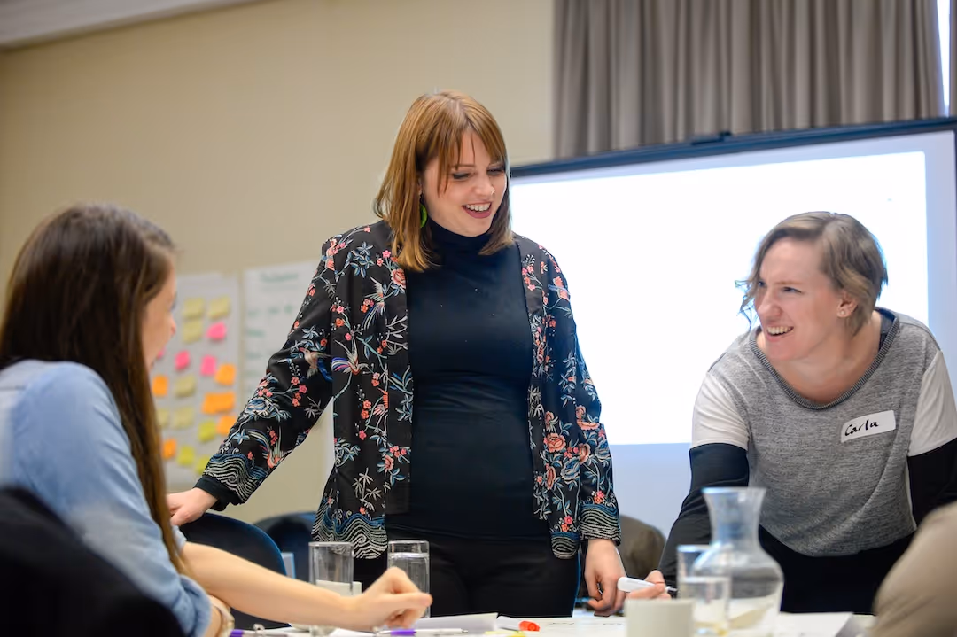 Three women laughing and collaborating at a table with notes and glasses in a meeting room.
