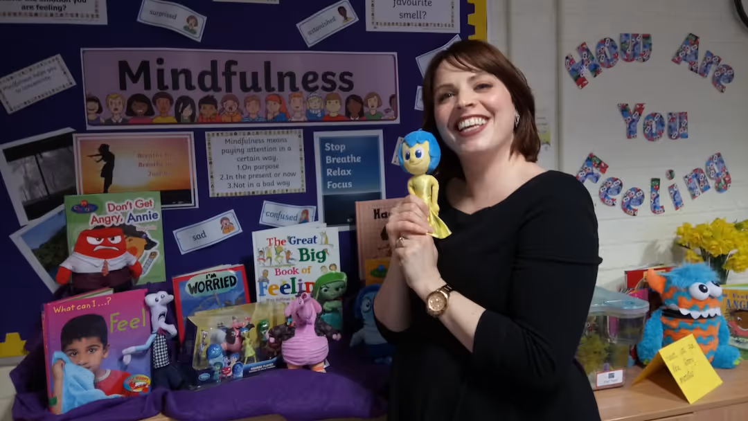 Liz Robson in black holding blue-headed doll with mindfulness books and mood posters behind her.