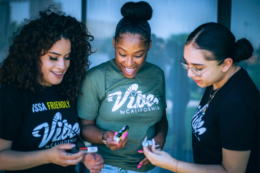 Three women from Vibe by California smiling and examining small items in their hands outside a glass building.