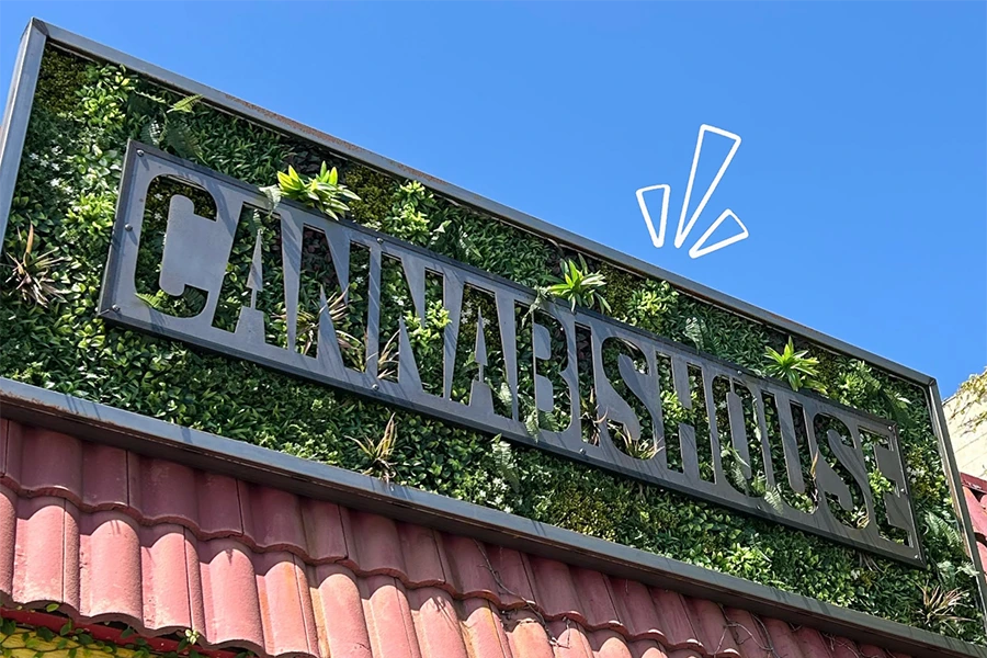 Metal sign reading CANNABIS HOUSE on a green leafy wall above a red tile roof under clear blue sky.