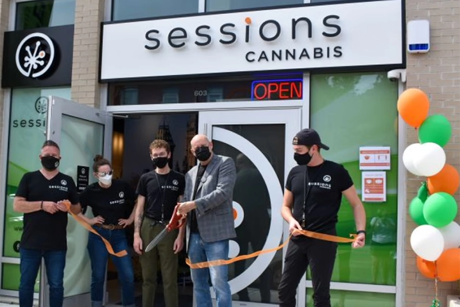 Group wearing masks cutting ribbon in front of Sessions Cannabis storefront with orange, green, and white balloons.