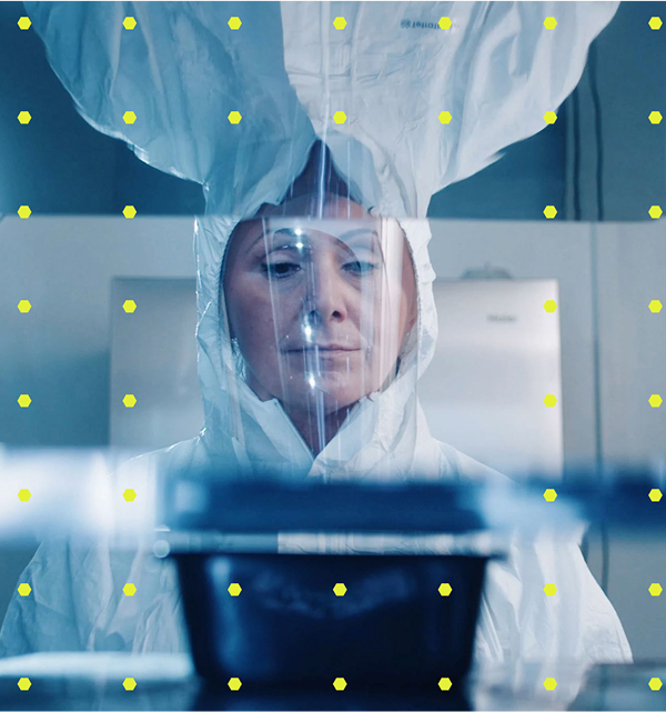 A picture of a woman in a lab wearing protective material, working on a machine. There are yellow dots placed over the image.