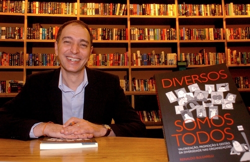 Imagem de um homem branco vestido de roupa social e sorrindo. Ao fundo da foto, uma estante repleta de livros, a direita do homem tem um livro com o título: "Diversos somos todos"