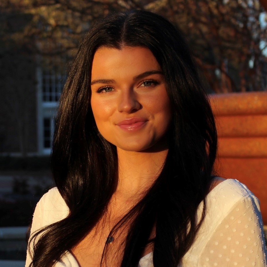 A woman with long, straight, black hair smiling at the camera. The picture is taken outside with a tree in the background.