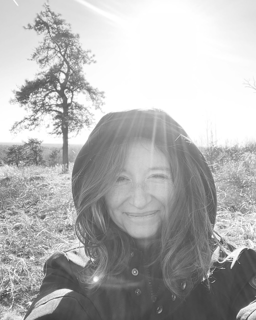 Black and white photo of a middle aged white woman with long hair wearing a black coat, standing in front of a tree. 