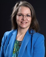 The photo is of a white woman with shoulder-length brown hair. She is wearing wire-rimmed glasses, a green and blue print blouse and a royal blue jacket. The background of the photo is dark. 