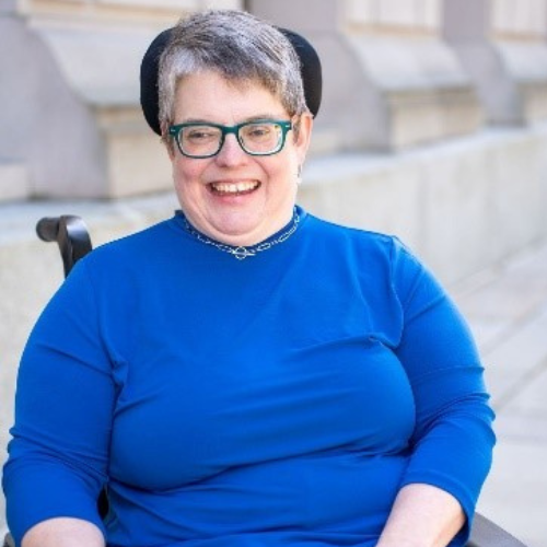 The photo is of a smiling woman who has short gray hair. She is using a wheelchair and her hand is on the controls. She is wearing green rimmed glasses, a royal blue sweater and pants striped in varying shades of blue. 