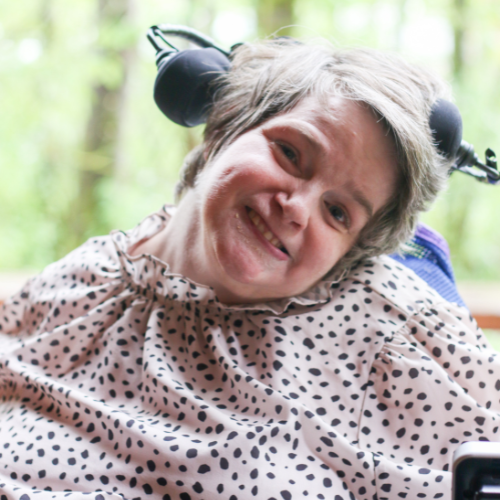 The photo is of a white woman with light colored short hair. She is sitting in a wheelchair with her head supported. She is wearing a pink blouse with black dots. She is pictured against a background of green trees. 