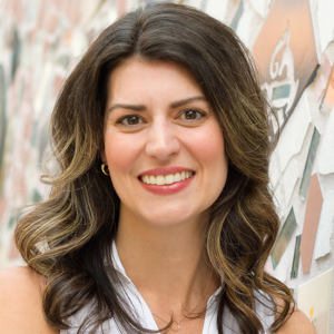 The photo is of a young white woman with long brown hair. She is smiling into the camera and wearing a sleeveless print blouse. 