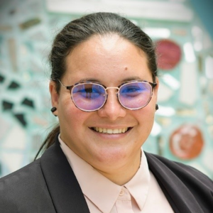 The photo is of a Latina person with hair pulled back. They are wearing glasses with light-colored frames, a tan shirt and a brown jacket, against a colorful background. 
