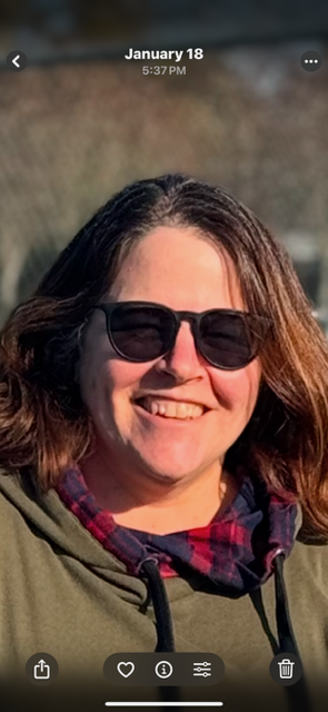 The photo is of a white woman in her late 50’s, with brown and grey streaked hair and hazel eyes. She is smiling into the sun, wearing sunglasses and an olive green jacket with a red plaid collar.