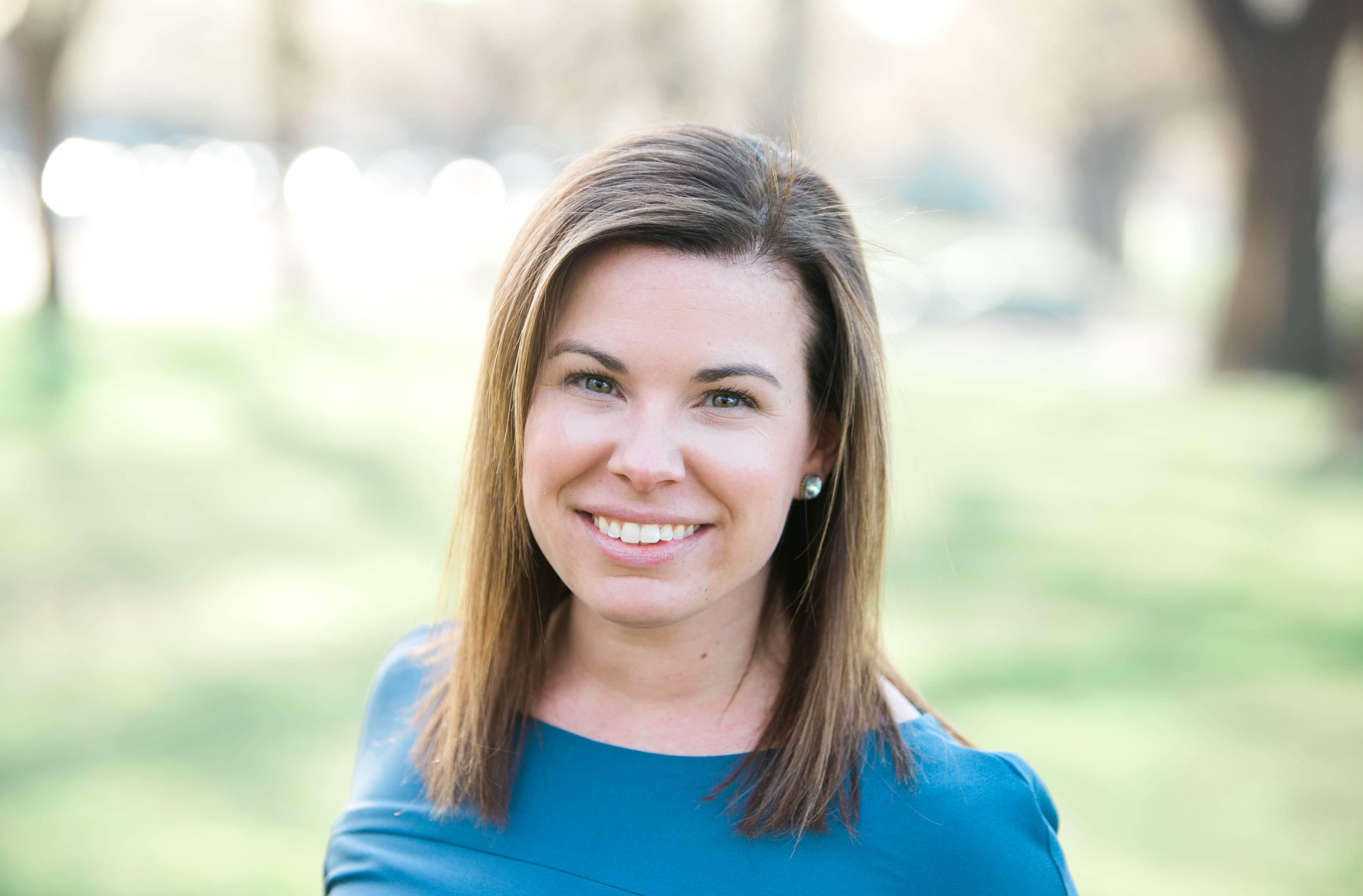 The photo is of a white woman with light-colored shoulder-length hair smiling into the camera. She is wearing a blue sweater and is against a background of trees. 
