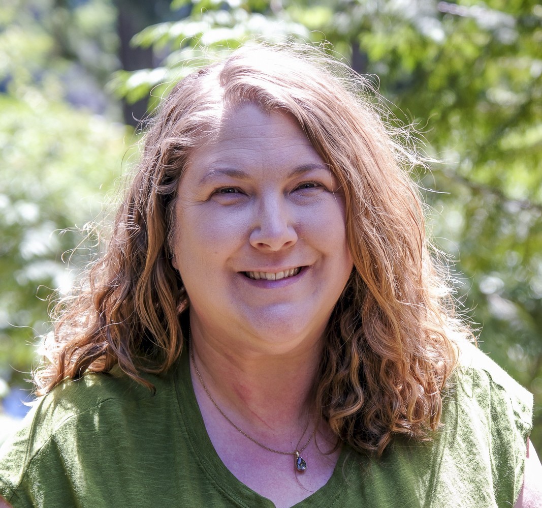 The photos is of a female with shoulder length blond hair and blue eyes, wearing a green shirt with a topaz necklace. In the background there are trees and sunlight. 
