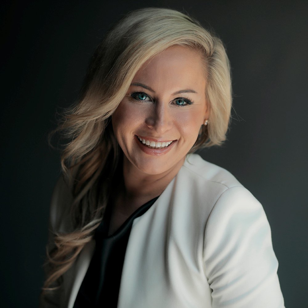 A woman with long, wavy, blonde hair smiling at the camera. She has on a white blazer.