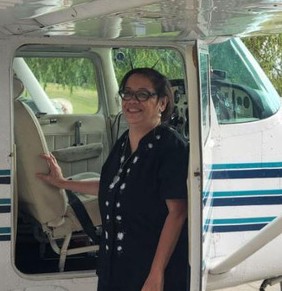 A woman with a black shirt and glasses, hair pulled back, stands in front of the cockpit of a small plane, smiling at the camera.