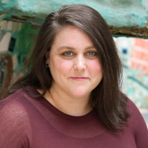 A woman with dark shoulder length hair wearing a maroon shirt against a blue background