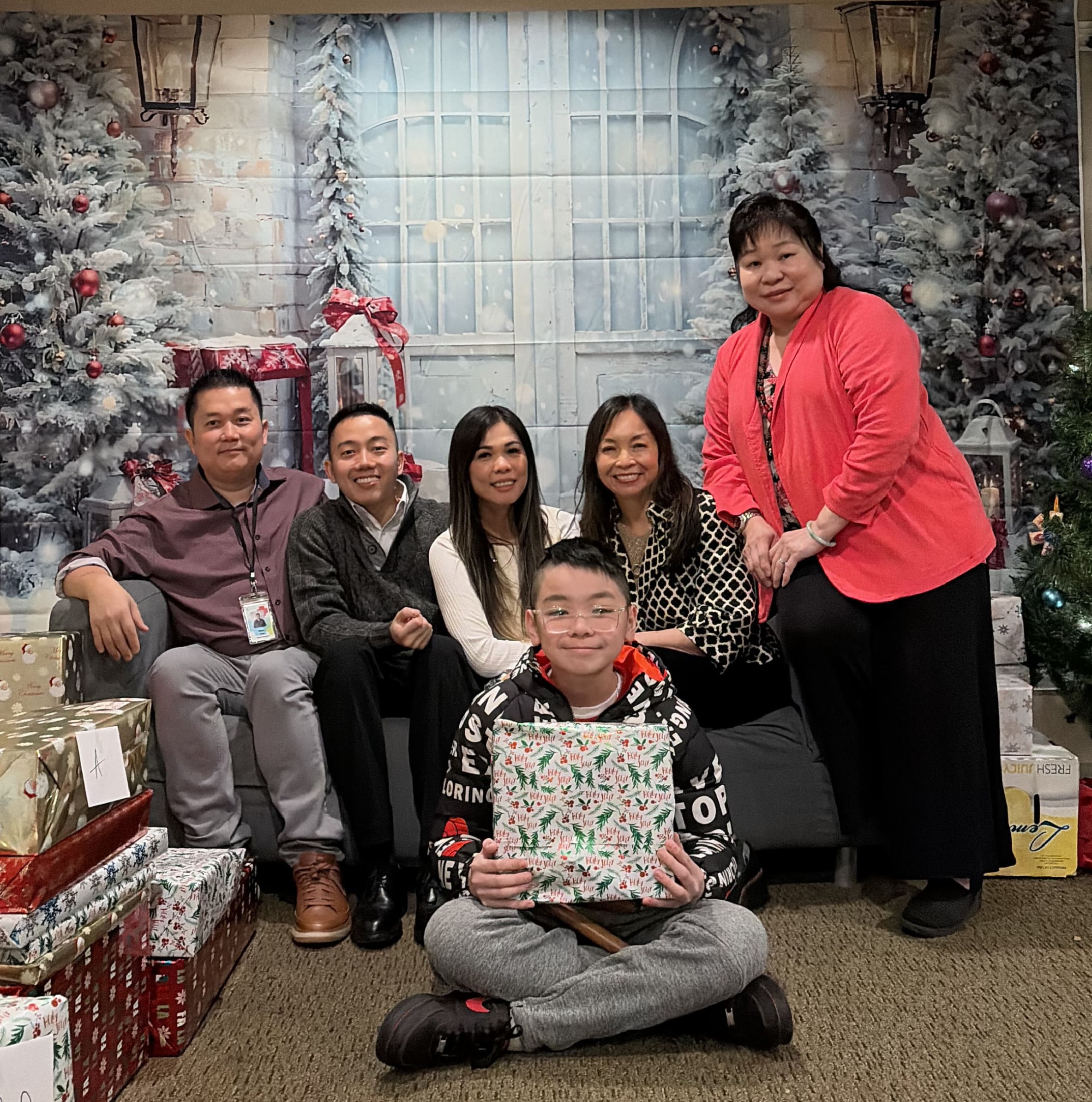 Six adults sit together on a couch behind a child holding a wrapped gift, posing in front of a holiday backdrop with a Christmas tree and stacks of presents.