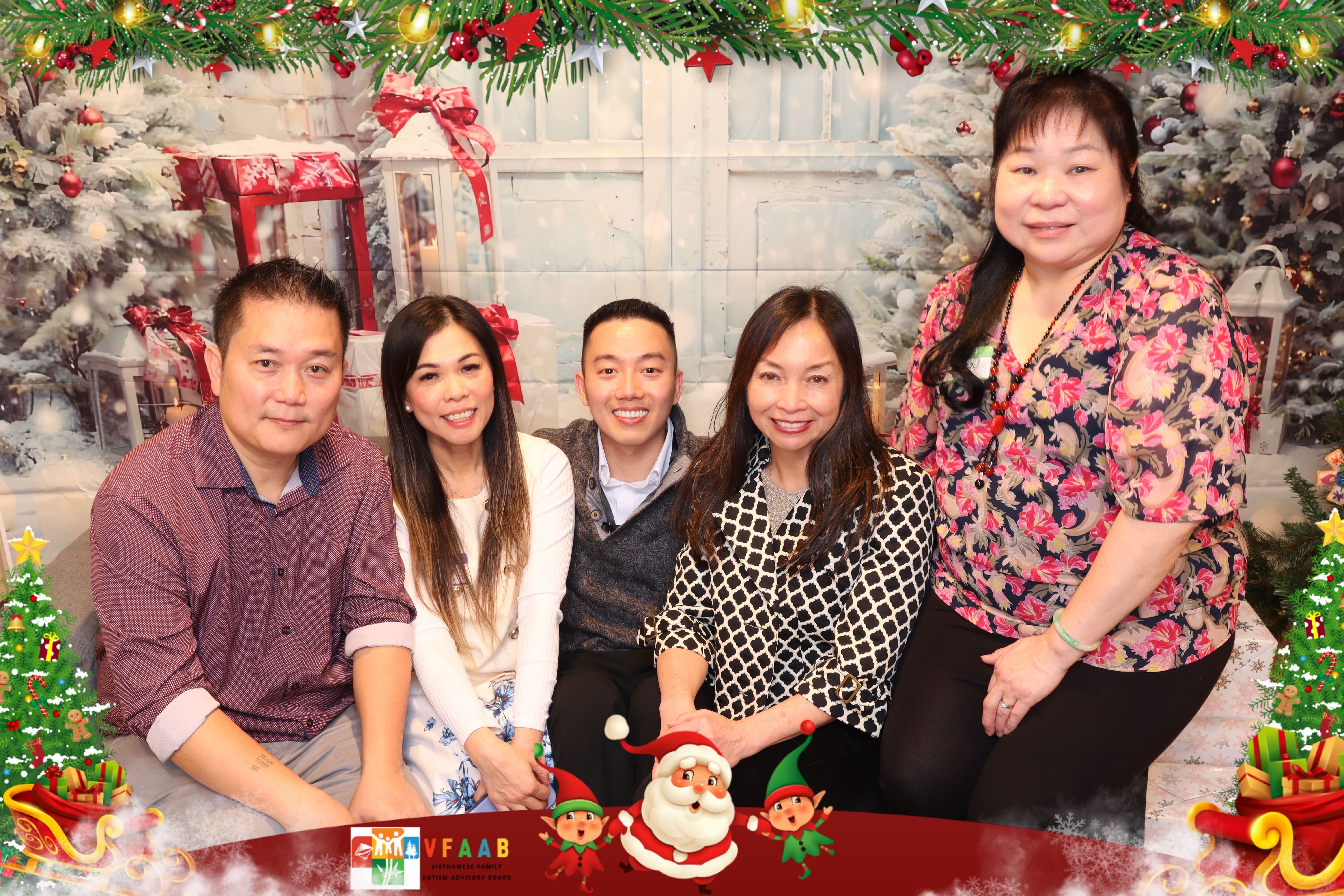 Five adults pose together in front of a festive holiday backdrop with greenery, lights, and wrapped gifts, smiling at the camera.