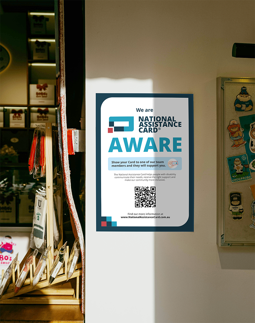 A poster on a shop wall promoting the National Assistance Card. The poster reads “We are National Assistance Card Aware.” It explains that customers can show their Card to staff to receive support. A QR code is displayed in the centre, with a link at the bottom to www.NationalAssistanceCard.com.au. The poster is framed by sunlight and surrounded by shelves of small gift items.