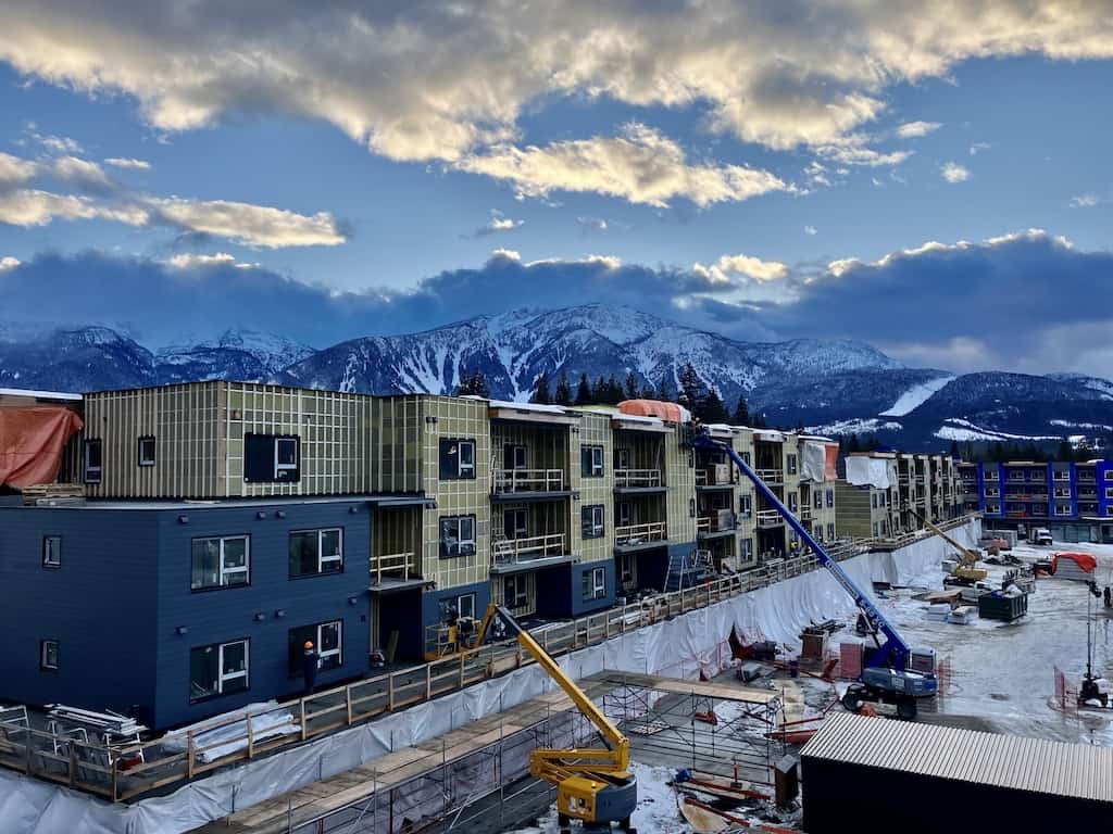 Dusk falls on a construction site with a partially completed building, a snowy mountain range in the backdrop, and construction equipment in the foreground.